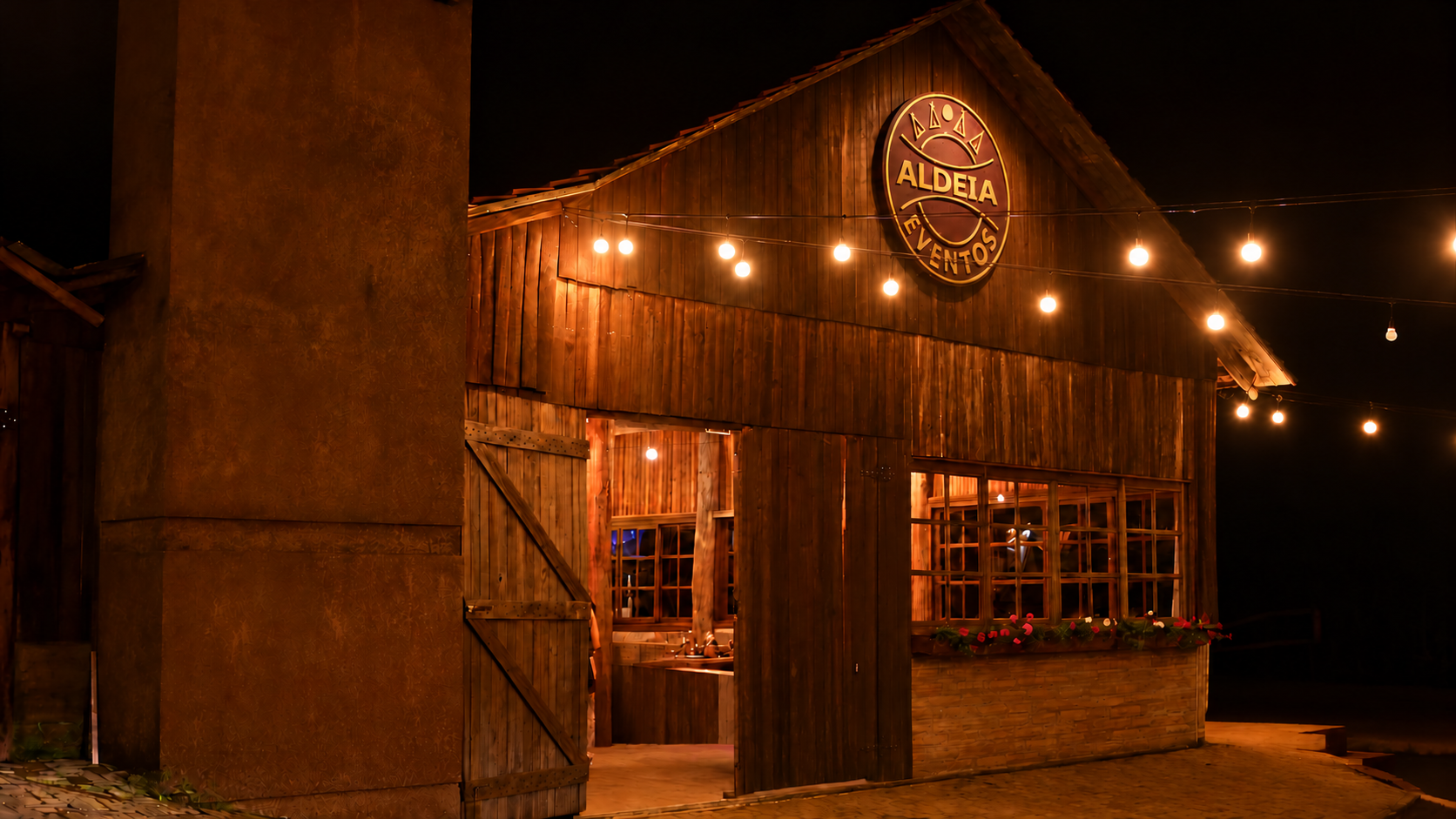 Fachada noturna da Aldeia Eventos, com brasão iluminado e entrada aberta